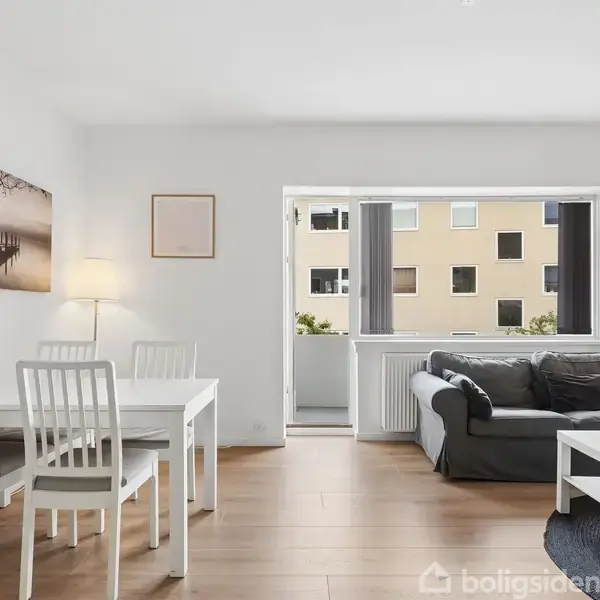 Et hvidt spisebord med fire stole står i et lyst stueområde. Til højre ses en grå sofa med et lille bord foran. Store vinduer mod en bygning udenfor.
