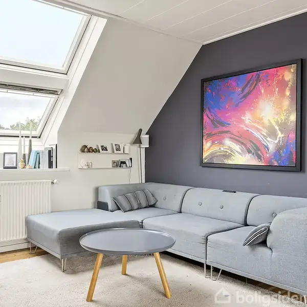Grå sofa i hjørne af lys stue, dekoreret med farverigt abstrakt maleri på en mørkegrå væg. Store vinduer med udsigt, rundt sofabord og hylder med billeder.