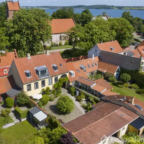 Et gult hus med rødt tegltag og store vinduer er omgivet af en frodig have. Omkringliggende bygninger har lignende arkitektur. Skoven og havet ses i baggrunden.