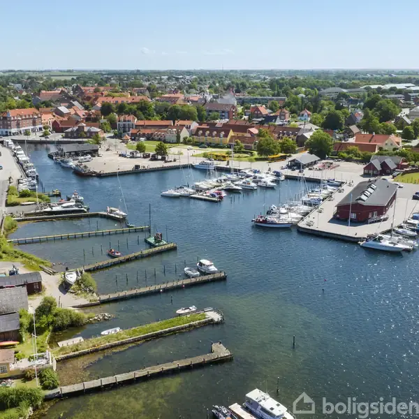 En havn med flere fortøjede både ligger ved siden af en by. Omkringliggende bygninger, veje og grønne områder omgiver havneområdet, med en klar blå himmel og frodig vegetation i baggrunden.