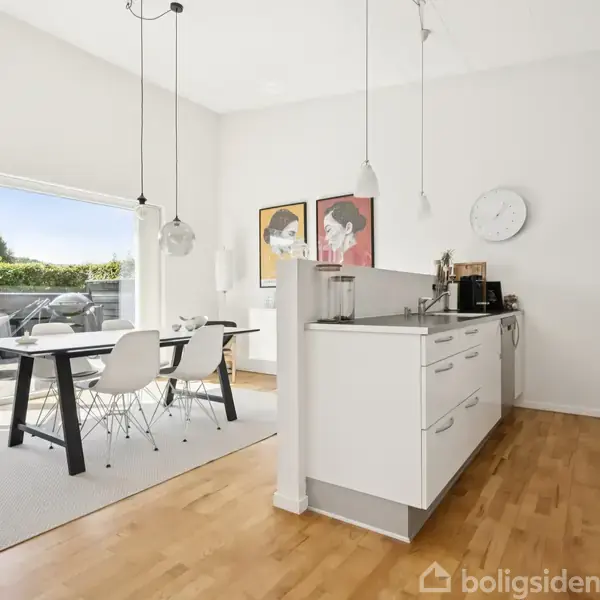 Et lyst køkken-alrum med hvide skabe og træparketgulv. Et spisebord med stole står ved glasdøre, der fører til en terrasse. To moderne billeder hænger på væggen.