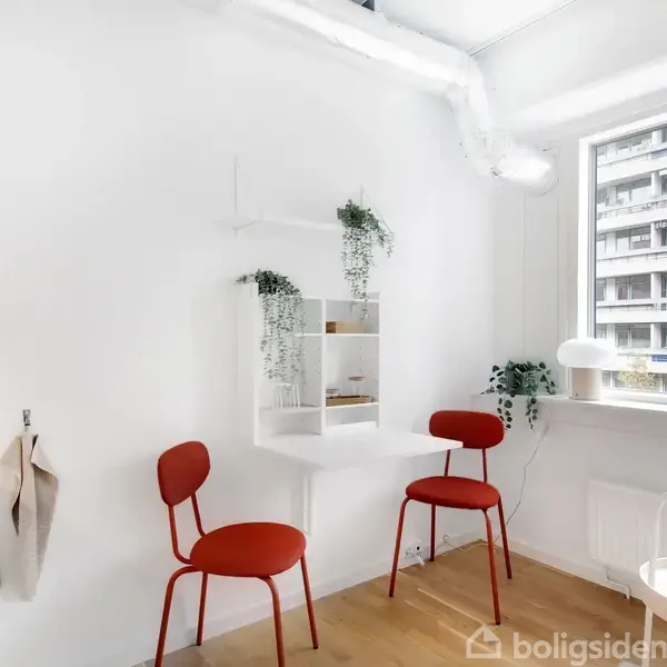 To røde stole står i et lyst rum med hvide vægge. En lille hvid hylde med hængende planter hænger på væggen. Store vinduer giver udsyn til en bygning udenfor.