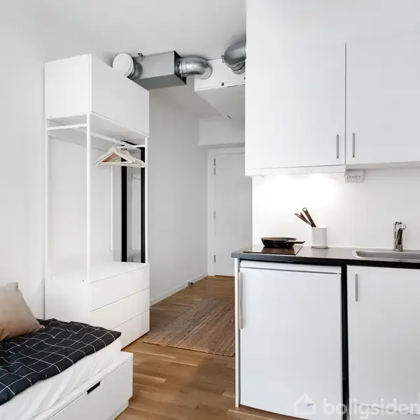 Et lille køkken med vask og skabsplads vises ved siden af en seng med puder. Til venstre er et åbent garderobeskab. Ovenover hænger en plakat med teksten "Merci New York."