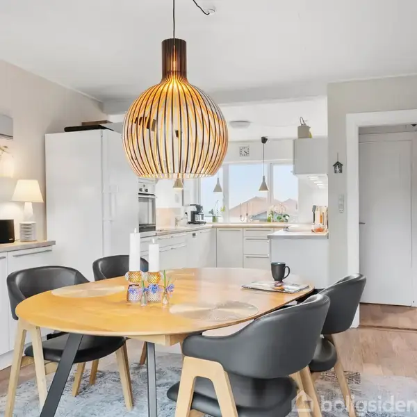 Rundt spisebord med fire sorte stole står på et tæppe. Over bordet hænger en trælampe. Baggrunden viser et moderne køkken med hvide skabe og dekorationer.