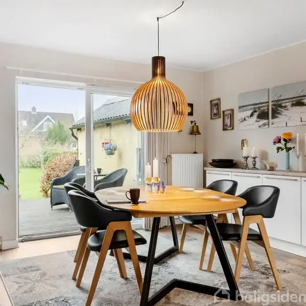Spisebord med fire sorte stole står i en lys spisestue med trægulv. Bordet er dækket til to. En stor moderne lampe hænger over bordet. Glaspaneldør åbner til have.