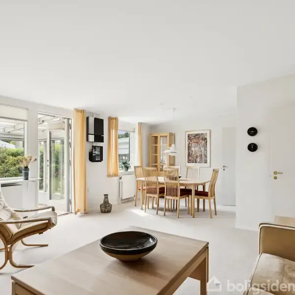 Et elegant stueområde med lænestol og sofabord. Spisebord med stole står tæt ved store vinduer, der slipper lys ind. Billedkunst pryder væggene, og gule gardiner hænger ved vinduerne.