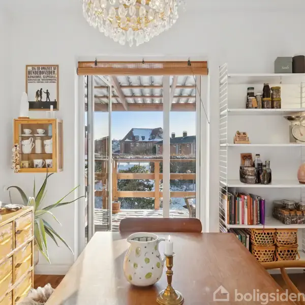 Et træbord står i en spisestue med en vase og en lysestage på. Hylder med bøger og krukker er på højre væg. En glasdør leder ud til en terrasse med udsigt.