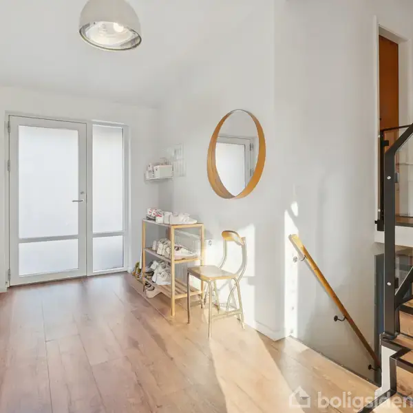 En lys entré med træparketgulv har jakker og hatte hængende på venstre væg. Et rundt spejl hænger til højre over en stol og en skohylde. En trappe fører opad.