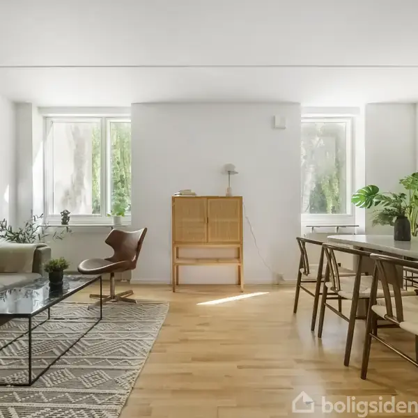 Sølvgrå sofa står ved siden af et lavt sofabord med glasplade på et tæppe med mønster. Ved siden af er et spisebord med otte stole. Rummet har lyse vægge og gulve.