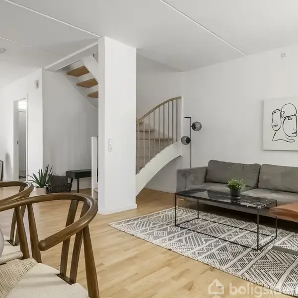 En stue med en grå sofa og et brunt læderstol ved et sofabord. Over sofaen hænger et minimalistisk kunstværk. I baggrunden ses en trappe og et spisebord.