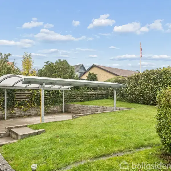 En overdækket terrasse med et gennemsigtigt tag står i en velplejet have. Græsplænen er omgivet af tætklippede hække og forskellige buske. Skylinen viser enkelte skyer på en blå himmel.