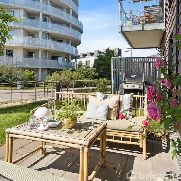 Terrassemøbler står på fliser ved siden af en lille blomstrende rosebusk. I baggrunden ses en moderne lejlighedsbygning og en grill, med træer langs vejen i et urbant miljø.