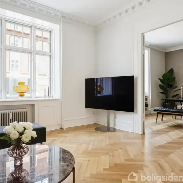 Stue med en vægmonteret fladskærms-tv, blomster i vase på sofabordet, store vinduer til venstre, og spisestue synlig gennem en døråbning til højre i rummet.