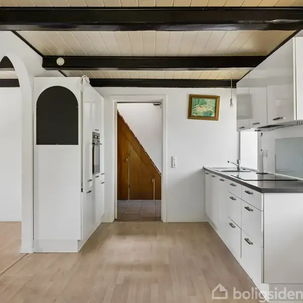 Køkken med hvide skabe udfører ingen handling, men rummet viser en åben indgang til en gang med trætrappe. Sort loftsbjælker og et maleri på væggen skaber hygge.