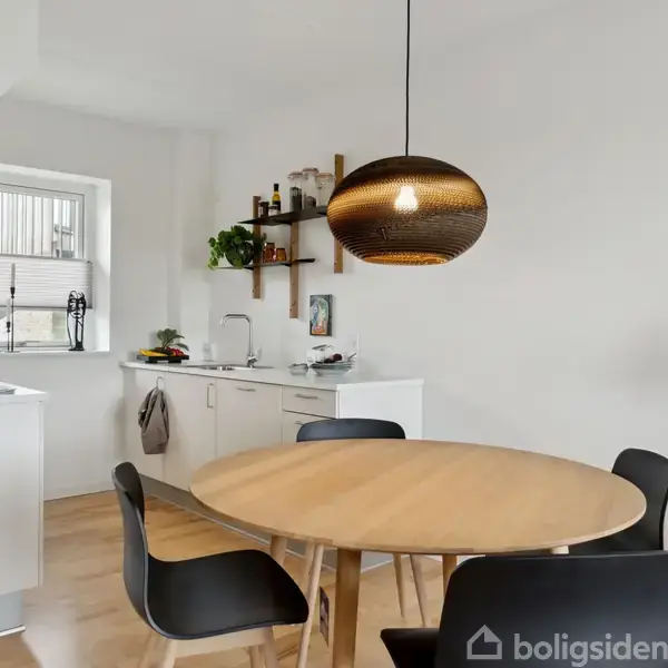 Rundt spisebord med sorte stole står under en hængende lampe i et moderne køkken. Hvide skabe og en vask fylder rummet, og der er planter og flasker som dekoration.