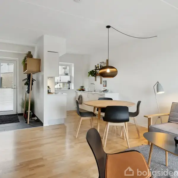 Rummelig stue med træbord og stole under en hængelampe, åben forbindelse til køkkenet, dekorativt sofabord, sofa med farverige puder og planterne indrammer indgangen i en moderne lejlighed.