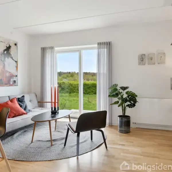 Sofa med farvede puder står ved et rundt sofabord; glasdøre i baggrunden giver udsyn til en grøn have. Stuen er dekoreret med planter, kunst og lysindfald fra vinduet.