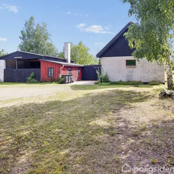 Et hus med sort tag og røde vægge står i en have med træer og græsplæne. Ved siden af huset er der en åben forplads og en hvid skorsten.