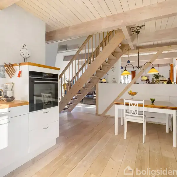 En moderne, lys køkkenet med træelementer, hvor et spisebord med stole står midt i rummet. Trappen fører til en øvre etage, mens hylder og køkkenredskaber pryder væggene.