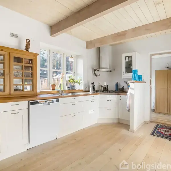 Køkken med hvide skabe og træbordplade, indeholder glasdørskab og opvaskemaskine. Solskin gennem vinduer belyser rummet, som har lyse trægulve og synlige loftsbjælker. Der er blå vaser på hylden.