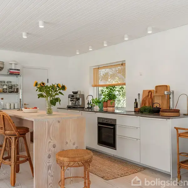 Køkken med en hvid ø og tre fletstole. Øen har en vase med solsikker. Det moderne rum har åbne hylder, hvide skabe, og køkkenmaskiner ved vinduet.