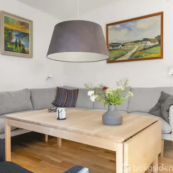 Grå sofa med farverige puder står langs en væg med malerier. Træbord med vase og blomster foran. Store, moderne loftlampe hænger over bordet. Trægulv og lys vægfarve.