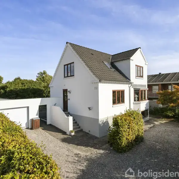 Et hvidt hus med mørkt tag står på en grusvej omgivet af grønne buske. Nummeret "23" ses ved hoveddøren. Omkring ligger en have med træer og en garage.