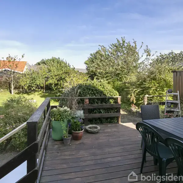 Træterrasse med havemøbler og potteplanter står i solrigt vejr. Terrasse omringet af grøn have med træer og buske; hus med rødt tag kan ses i baggrunden.