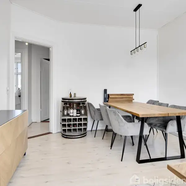 Spisebord i træ med grå stole omkring, står i moderne spisestue med lyse vægge og gulv. En sort og træfarvet skænk til venstre og vinreol foran en åbning til en lys gang.