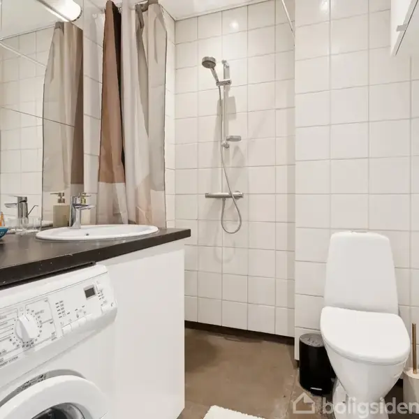 Vaskemaskine på badeværelse ved siden af håndvask med spejl, bruser i hjørne, toilet ved siden af lille skraldespand. Væggene er beklædt med hvide fliser.