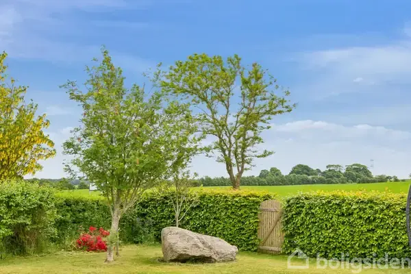 En stor sten ligger i en grøn have med to træer og en busk med røde blomster. I baggrunden ses en hæk, en træport og en åbning mod marker.