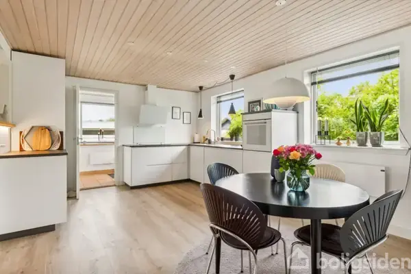 Et rundt spisebord med blomster i en vase står i et lyst køkken med hvide skabe, store vinduer og trælofter. Grønne planter står på vindueskarmen.