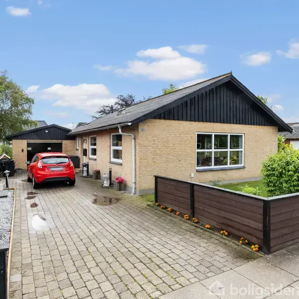 En rød bil holder parkeret på en flisebelagt indkørsel foran et gult murstenshus. Huset har et sort gavltag, og omgivelserne inkluderer hække og et træ.