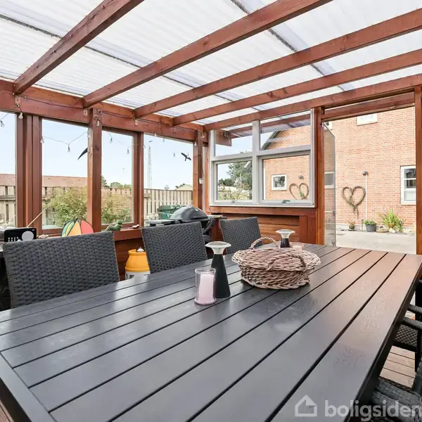 Sort bord med kurvestole omkring står på en træterrasse i et overdækket område med glasvægge. Murstenshus og trægærde ses i baggrunden. Lyse lyskæder hænger over bordet.