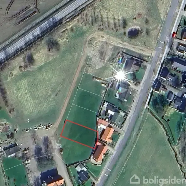 Luftfoto viser et boligområde med flere huse langs en vej. En ledig grund på en grøn mark er markeret med en rød kant. Omkringliggende landskab består af marker og spredte træer.