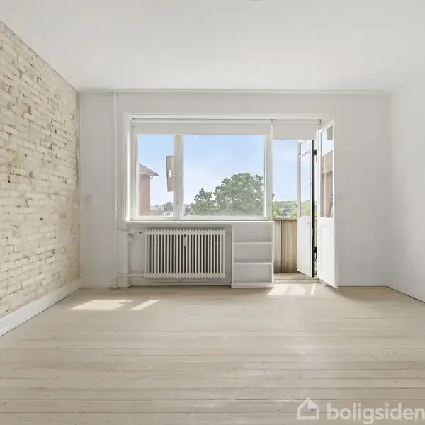 Radiator under vindue med åben dør til balkon. Rød stenbygning ses udenfor, omgivet af træer og blå himmel.