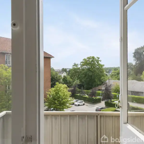 Træer og buske står i en frodig have omgivet af en muret bygning med flere altaner. Græsplænen er velplejet, og der er blomstrende planter rundt om i haven.