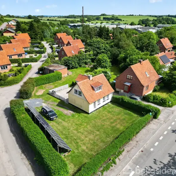 Røde tage af huse i en beboelseskvarter. En bil parkeret ved siden af et hus omkranset af hække. Træer og græsplæner omgiver huse i en grøn, frodig landsby.