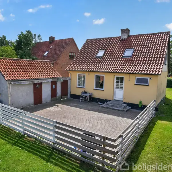Et gult hus med rødt tegltag står ved en flisebelagt gårdhave omgivet af et hvidt hegn. Ved siden af er en garage. Baggrunden er græsplæne og blå himmel.