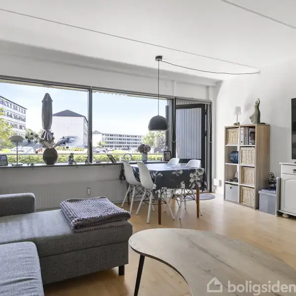 En lys stue med en grå sofa og træbord foran. Et spisebord med stole står ved et stort vindue, der viser en udsigt over en bygning. En stor fladskærm er placeret på en hvid skænk til højre. Reoler med bøger og dekorationsgenstande står ved siden af.