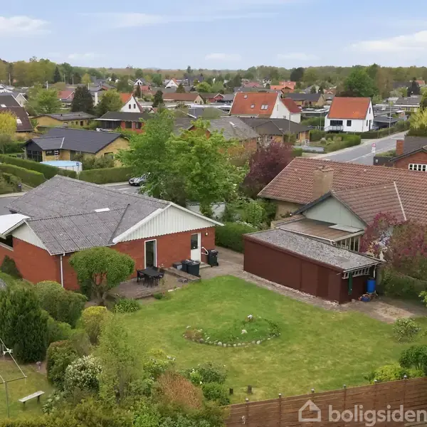 Huset i forgrunden er rødt med en grå tag. Haven har græs og buske. Det ligger i et boligområde med flere lignende huse, omgivet af veje og træer.
