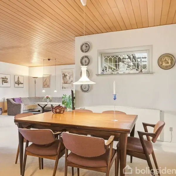 Et spisebord med fire stole står i et lysindfald fra en moderne lampe, omgivet af hvide vægge dekoreret med kunst og et vitrineskab i baggrunden.