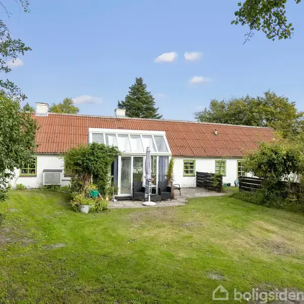 Hus med rødt tegltag har en terrasse med glasparti. Græsplæne og træer omkranser huset i en stille, landlig have.