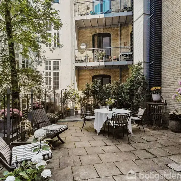 Terrasse med bord og stole, omgivet af grønne planter og blomster i krukker. Baggrunden består af høje, smukke murstensbygninger med altaner og store vinduer. En afslappende udendørs oase.