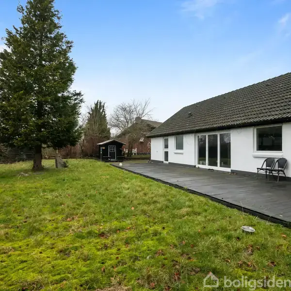 Hus med hvid facade og sort tag står ved siden af græsplæne og terrasse med bord og stole. Træ og skur i baggrunden under blå himmel.