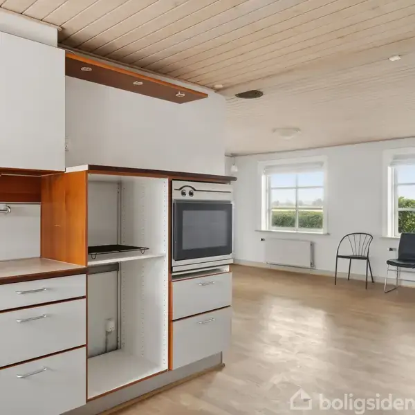 En moderne køkkenenhed med integreret ovn og skuffer står i en lys stue. Rummet har trægulve, et lyst træloft og store vinduer, hvor der ses udsigt til grønne omgivelser.