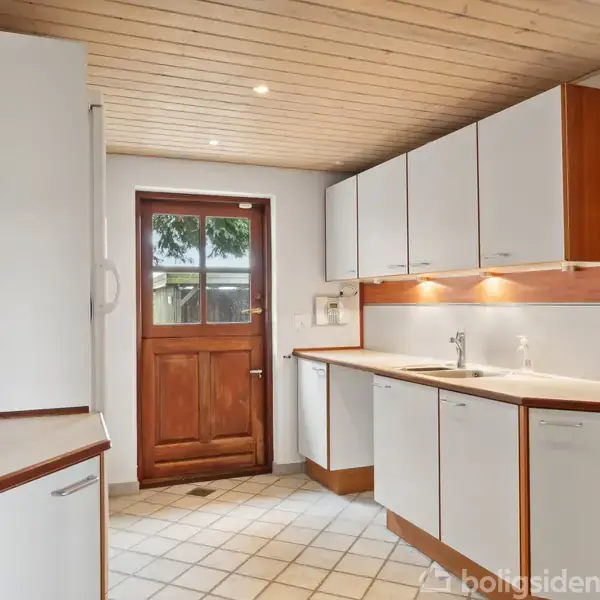 Et køkken med hvide skabe og en trædør. Skabene er indbyggede, med en vask og underskabsbelysning. Flisegulv og træloft med indbyggede spots.