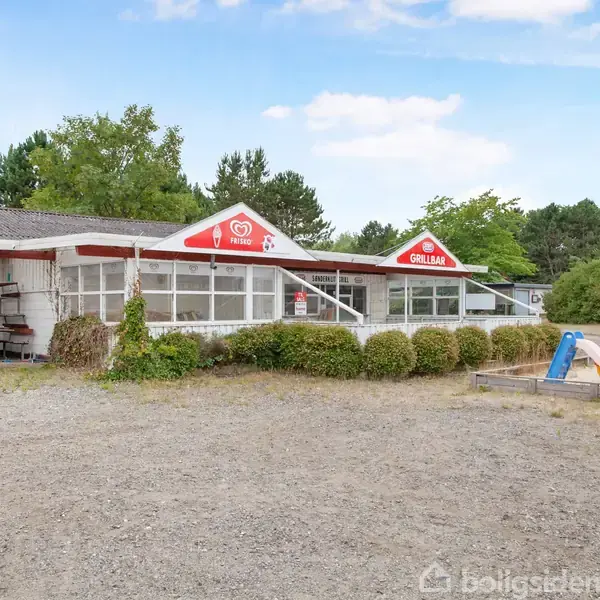 En isbutik med to store røde "Cornetto" og "Grillbar" skilte står i et grusbelagt område. Mindre buske grænser op til bygningen, og en legeplads ses til højre.