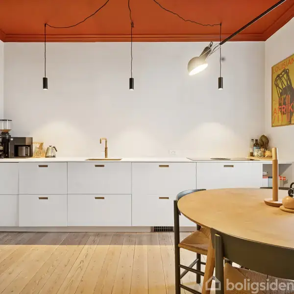 Et lyst køkken med hvide skabe og trægulv. En rund træbord står ved et vindue, og en plakat med teksten "Visitez L'Afrique" hænger på væggen.