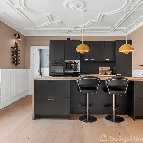 Et moderne køkken med sorte skabe og to barstole. Over bordpladen hænger to gule lamper. Store vinduer og dekorativt loftpanel i et lyst rum. En vinkaraffel ses i vindueskarmen.